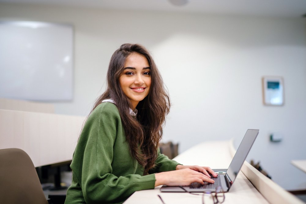 Uma estudante universitária em sala de aula, sorrindo para câmera e mexendo no notebook.